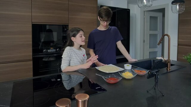 Brother and sister are preparing a pizza by recording a video with their smartphone in a modern kitchen, sharing their culinary skills with online audience
