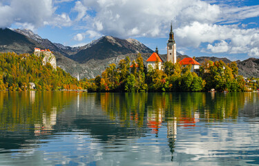 Lake Bled on a sunny autumn day. A view of the island and the Church of the Assumption of Mary. Traveling through Europe, Slovenia. A forest on the cliffs on a sunny day. Tourist attractions of Europe