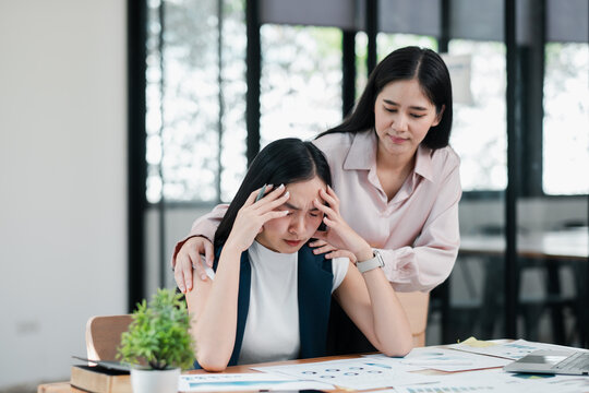 A caring colleague consoles a stressed businesswoman at her desk in a bright, modern office setting.