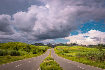 A low-angle shot of an open country road with lush green scenery, dramatic clouds, and a blue sky.
