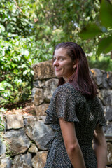 Fototapeta premium A smiling woman in a floral dress stands outdoors in front of stone steps surrounded by lush greenery. The natural light and vibrant backdrop enhance the cheerful and serene atmosphere