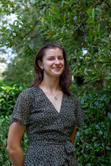 A smiling woman in a floral dress stands outdoors in front of stone steps surrounded by lush greenery. The natural light and vibrant backdrop enhance the cheerful and serene atmosphere