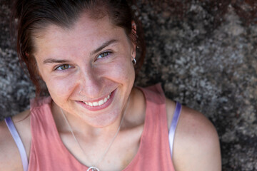 A close-up portrait of a smiling young woman with a nose ring, wearing a pink top. Her expressive eyes and radiant smile stand out against the textured rock background, creating a natural, candid vibe