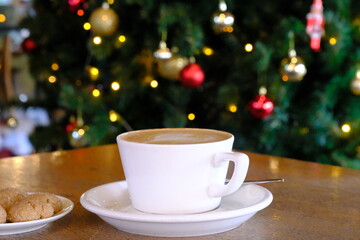 New Year's pine tree in the background. Cafe latte with a heart on it in a white cup in a cafe. Cinnamon cookies on the side. Merry christmas. New year preparations.
