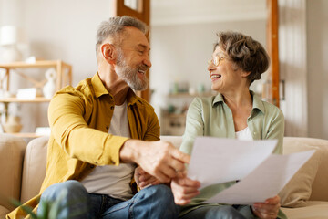 Positive European senior spouses checking and paying bills online, holding papers and banking on Internet, sitting on couch