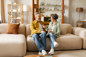 Happy senior couple talking, relaxing together on sofa at home, spouses enjoying spending time together, full length shot