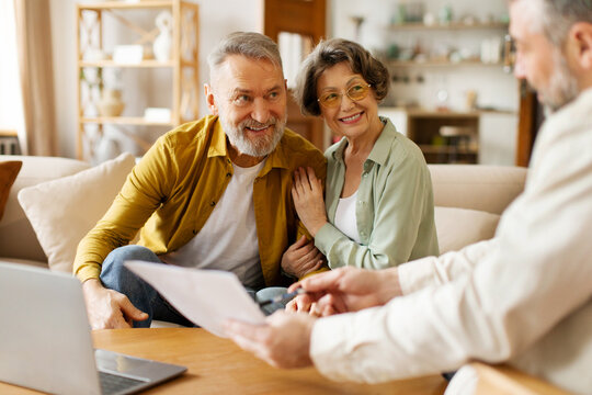 Realtor or broker showing documents and house plan to excited senior couple, agent or consultant help mature spouses with bank loan or mortgage at briefing