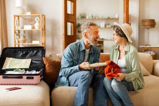 Happy senior couple packing clothes for trip together, sitting on sofa in living room and chatting, preparing for travel - Powered by Adobe