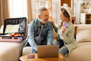 Happy senior spouses using laptop and booking hotel online with credit card, sitting with suitcases...