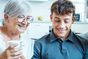 Elderly grandmother and young grandson sitting comfortably on a sofa in the living room, enjoying tea, smiling, laughing, and bonding while sharing cheerful stories during a festive family gathering
