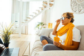 Adult woman relaxing on a sofa or chair in her cozy living room during the afternoon, holding a warm cup of tea or coffee, thinking about her future, and enjoying a peaceful and private moment