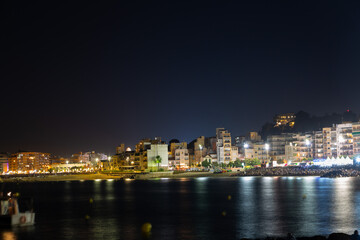 People relaxing on beach at night, watching summer festival with illuminated buildings in background