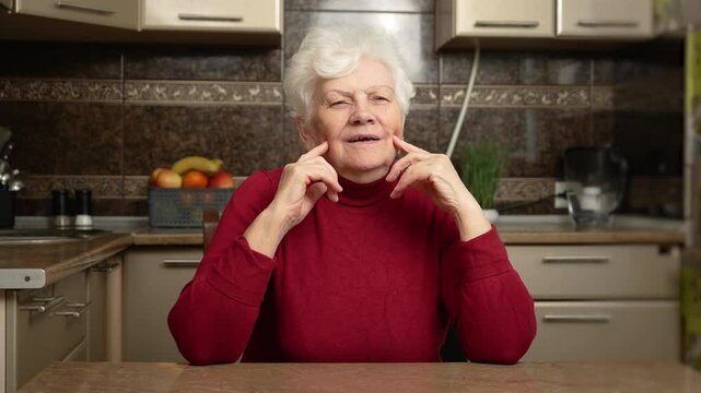 Elderly woman is being silly. Old female fills cheeks with air and presses fingers against them, puckering lips like trumpet and exhaling air from mouth in kitchen Flabby and wrinkled skin on senior