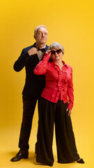 Sophisticated pair, man in tailored suit standing beside woman in modern attire and shades, posing confidently against yellow studio background. Concept of mature people in modern lifestyle, fashion.