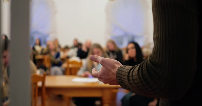 Man wearing brown turtleneck faces attentive crowd, delivering speech with purposeful intensity, emphasizing speech value in knowledge exchange, urging listeners to heed speech insights