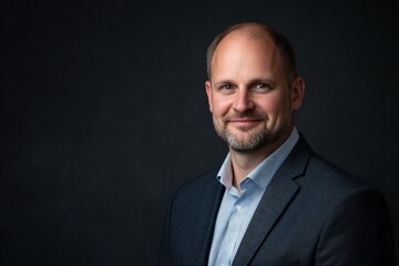 A confident middle-aged man with a short beard is wearing a blue suit and light blue shirt, posing against a dark background, emanating professionalism and warmth.