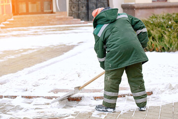 Woman in uniform with snow shovel cleans sidewalk in winter. Utility worker in uniform shoveling...