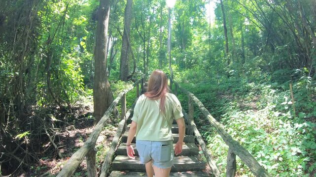 A young woman strolls along a wooden path in a vibrant green forest, surrounded by tall trees and lush foliage. The serene environment invites peace and reflection, perfect for nature lovers seeking t