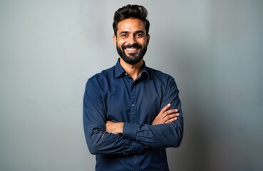 Confident Indian businessman stands in studio portrait. Successful entrepreneur corporate executive wears navy blue shirt. Beard. Arms crossed. Isolated against neutral gray background. Posing