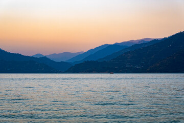 The blue lake features calm waters, with multiple layers of mountains in the background. Evening lights of the sun create a serene atmosphere, complemented by a small boat in the distance.