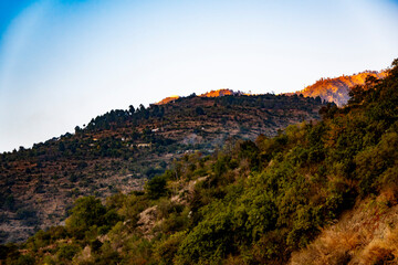 Golden sunlight bathes the mountain tops, illuminating small towns nestled in the hills. A full view of the mountains, with green cover, enhances the scenic beauty of Tehri Lake.