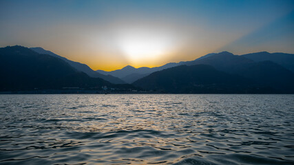 The sunset casts dramatic rays behind the mountains above the lake, with blue mountains and blue waters creating a stunning backdrop at Tehri Lake.