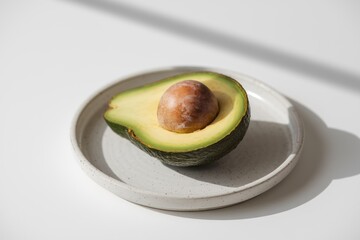 A halved avocado with a visible pit rests on a speckled plate, bathed in sunlight.