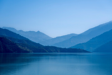 Multilayered blue mountains are shrouded in mist, with blue light cascading over them and the lake...