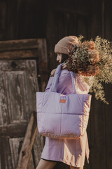 Naklejka premium A woman in a hat and a light purple jacket with a large quilted bag and a bouquet of dried flowers poses in front of a wooden wall in subdued light (selective focus)