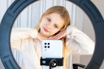 Young girl filming with ring light. Blonde woman using a smartphone and ring light to record video content, posing in a cozy white sweater indoors.