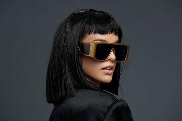 Blackhaired woman in sunglasses and black jacket posing confidently against a gray background