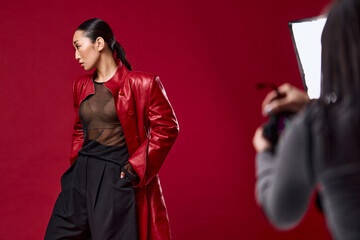 Fashion asian woman in red leather jacket and black pants posing for a photo with a photographer against a red background