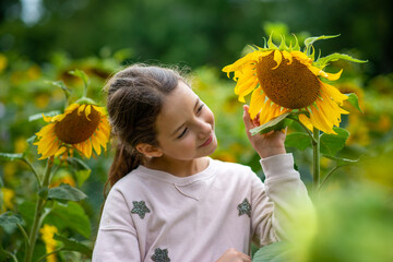 A young girl interacts joyfully with a large sunflower among an array of blooming flowers in a sunlit garden on a pleasant day.