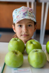 A young girl stands beside lush greenery, smiling cheerfully in an urban area. Her casual outfit and cap complement the sunny day atmosphere.
