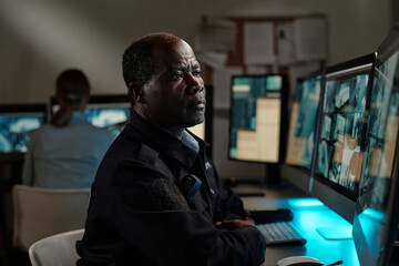 Serious African American security guard concentrating on CCTV video on screen of computer while...