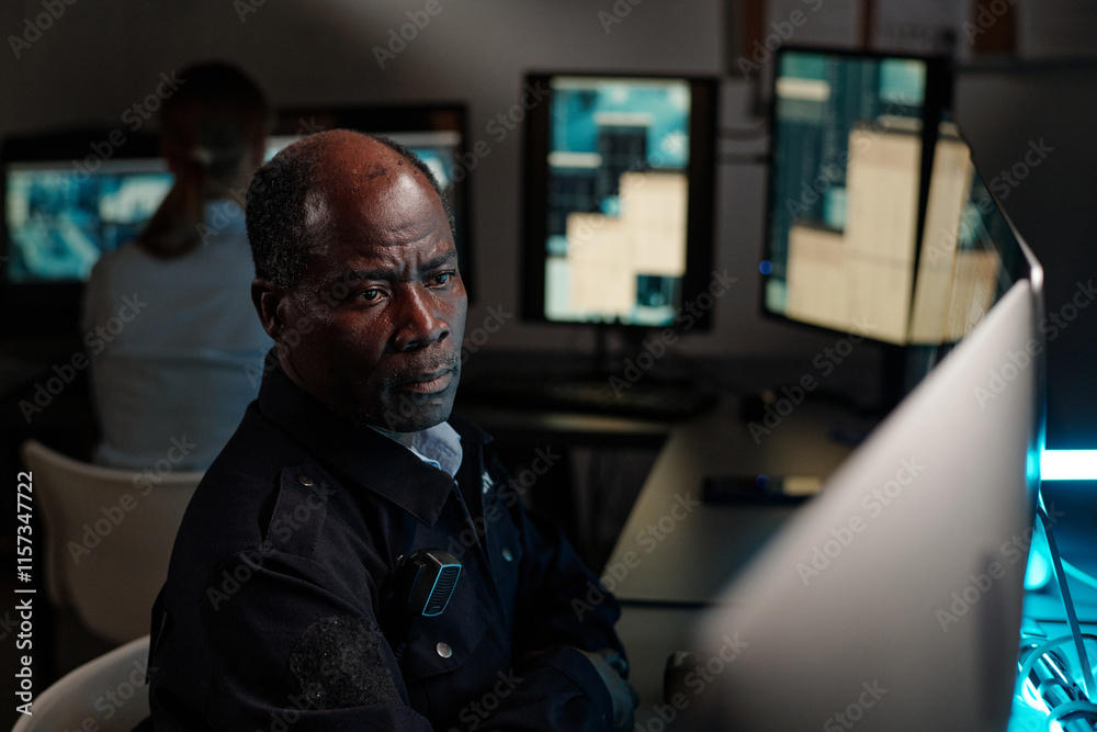 Poster serious mature security guard sitting by workplace in observation room and looking at one of desktop