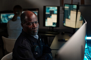 Serious mature security guard sitting by workplace in observation room and looking at one of...