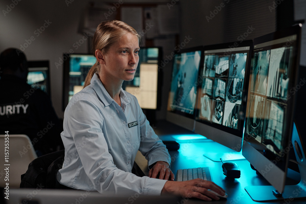 Poster young blond female security guard in uniform sitting by desk in control room, looking at desktop com