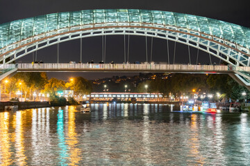 Night view of the illuminated pedestrian bridge over the river in Tbilisi with reflections and city...