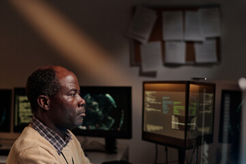 Serious mature man in beige sweater sitting in dark office and looking at computer screen while decoding online data