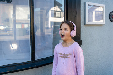 Singing joyfully with headphones on a sunny day. A girl wearing pink headphones passionately sings outside a charming building, embracing the joyful sounds of her favorite tunes under clear blue sky