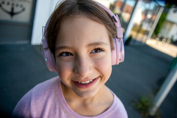 Joyful girl wearing pink headphones enjoys a sunny day outdoors. A cheerful young girl with brown hair is smiling brightly while wearing pink headphones.