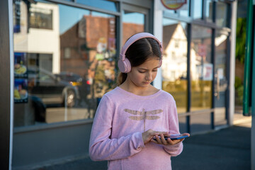 A young girl in a pink sweater enjoys music with headphones while standing outside a shop, focused on her phone and the vibrant street backdrop.
