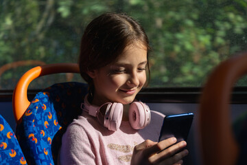 A young girl wears pink headphones while happily using her smartphone on a bus. Sunlight filters through the windows, illuminating her joyful expression.