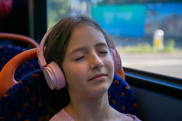 A young girl sits on a bus, lost in her thoughts, with headphones on. Her eyes are closed, indicating a serene escape as the city passes by outside.