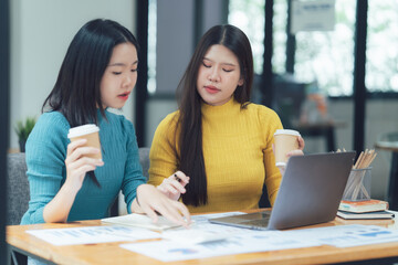 Collaborative Brainstorming: Two focused women, in a casual office setting, discuss ideas while reviewing documents and a laptop. Capturing the essence of teamwork and innovative thinking.