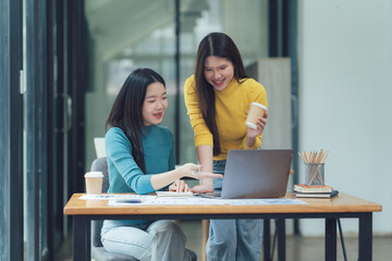 Collaborative Work Session: Two young Asian women engage in a productive work session at a modern office desk, using a laptop and discussing ideas.