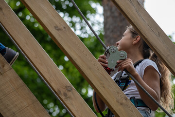 A young climber confidently ascends wooden beams while securely holding onto the safety equipment, surrounded by lush greenery in a vibrant outdoor park.