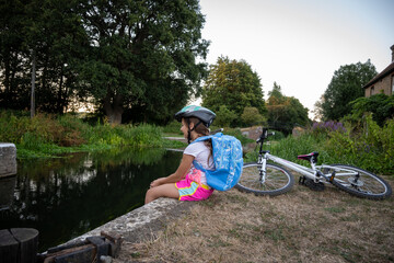 A young girl in a helmet rests by a calm waterway at dusk, enjoying the tranquility after exploring with her bicycle nearby, embraced by lush greenery.