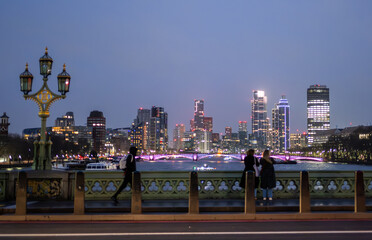 Nighttime view of London from Westminster Bridge, featuring the illuminated city skyline, Thames River, lit-up buildings, and people on the bridge. The sky is clear, emphasizing the city lights